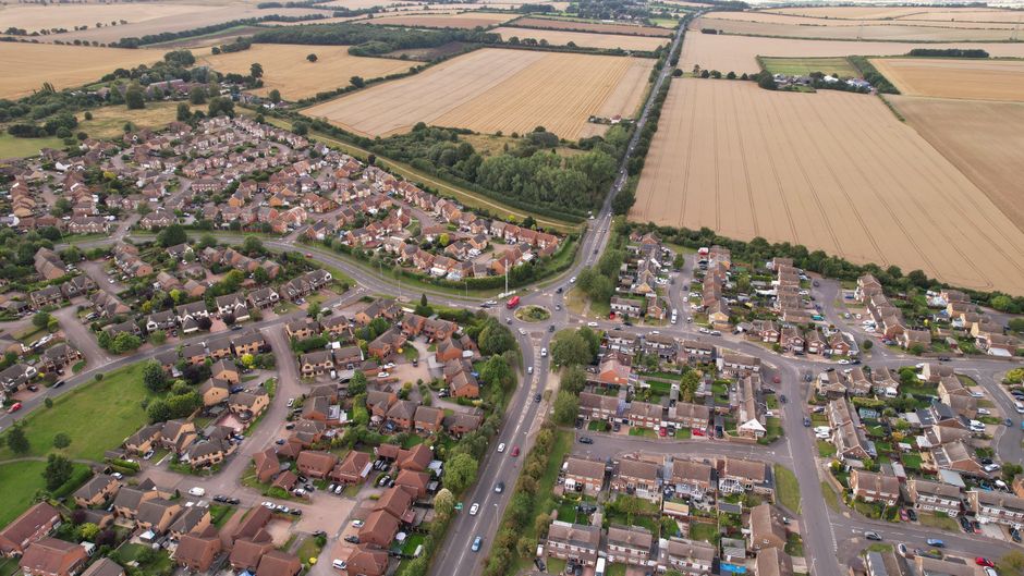 Aerial view of a residential neighbourhood with numerous detached and semi-detached houses, streets with parked cars, and lush green trees lining the roads. Surrounding the suburb are expansive farmla