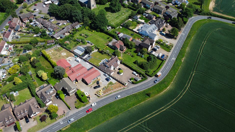 Aerial view of a residential neighbourhood with numerous detached and semi-detached houses, streets with parked cars, and lush green trees lining the roads. Surrounding the suburb are expansive farmla