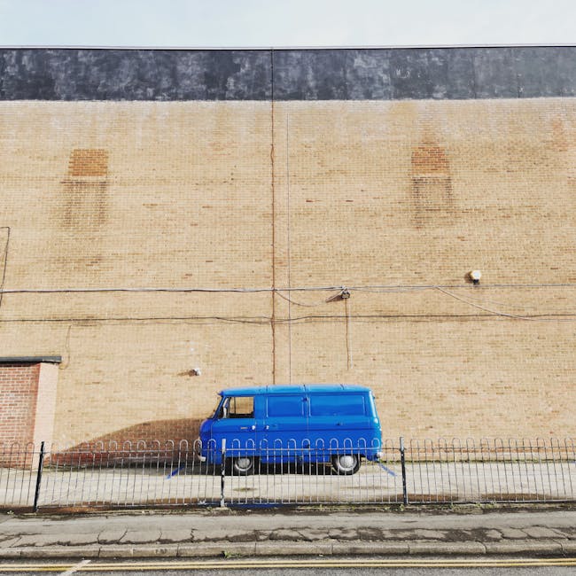 A blue vintage-style panel van parked on a paved sidewalk in front of a tall, beige brick wall with a black horizontal strip near the top. The van is positioned next to a black metal fence that runs parallel to the street. Overhead, there is a thin wire cable running across the wall, secured with small brackets and a visible padlock. The scene is outdoors in natural daylight, capturing the vehicle as part of a home relocation process, with the van likely used for furniture transport and packing during house removals and moving logistics, as indicated by the context of Beddington Lane flats and lift access advice on the associated webpage.