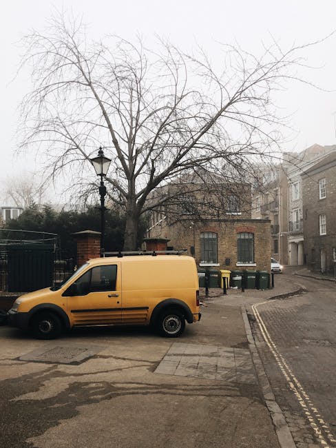 A yellow Man with Van Beddington van is parked on a residential street near a brick building with three arched windows fitted with black metal bars. The van is positioned close to the curb, partially on the pavement, with its side door closed. In the background, there are several green wheelie bins placed against the brick wall and a vintage-style street lamp nearby. A large, leafless tree is situated behind the van, extending above the building. The surrounding environment includes a narrow, slightly curved road with double yellow lines along the edge and a mix of traditional and modern houses in the distance. The scene appears to capture an early morning or overcast day, with diffuse natural light illuminating the area. This image relates to home relocation or furniture transport services as provided by Man with Van Beddington, illustrating the vehicle used for packing and moving belongings during house removals in the Beddington area.