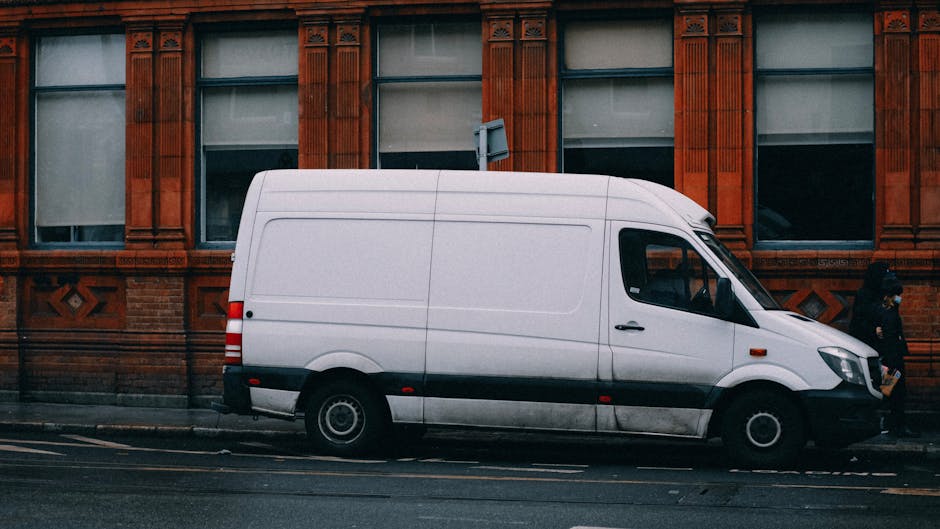 A blue vintage-style panel van parked on a paved sidewalk in front of a tall, beige brick wall with a black horizontal strip near the top. The van is positioned next to a black metal fence that runs parallel to the street. Overhead, there is a thin wire cable running across the wall, secured with small brackets and a visible padlock. The scene is outdoors in natural daylight, capturing the vehicle as part of a home relocation process, with the van likely used for furniture transport and packing during house removals and moving logistics, as indicated by the context of Beddington Lane flats and lift access advice on the associated webpage.