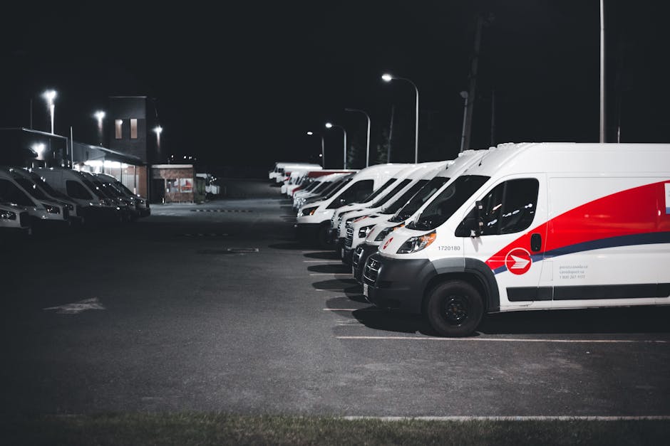 A row of white commercial vans with red and black branding parked in a well-lit, open parking lot during the night. The vans are positioned side by side, with their front ends facing the viewer, and t