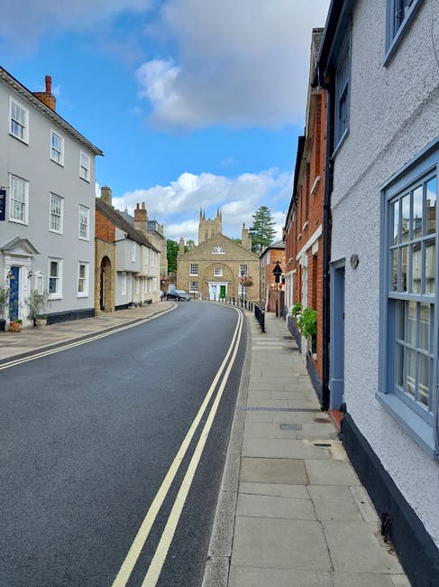A quiet residential street in Beddington with a gently curving asphalt road, lined by a mix of terraced and semi-detached houses with white, red brick, and painted facades. Some houses display potted plants near doorways and small front gardens. The pavement on the right side has evenly spaced concrete slabs, and a black metal bollard is visible along the sidewalk. In the background, a historic church with a prominent tower and battlements stands at the end of the street under a bright blue sky with scattered clouds. The scene is well-lit and appears to be daytime. This setting reflects a typical UK suburban area suitable for home relocation and furniture transport, often serviced by companies like Man with Van Beddington during moves involving packing and loading processes.