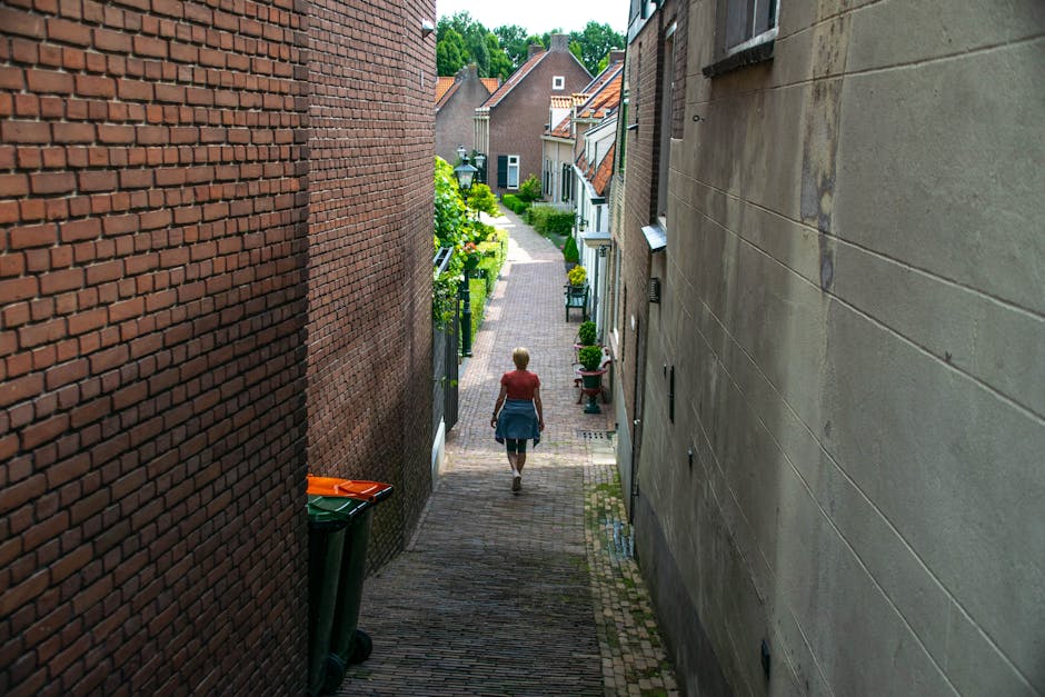 A narrow residential alleyway during daytime, with brick and concrete buildings on either side. A woman with blonde hair, wearing a red top and denim skirt, is walking away from the camera along a paved path lined with potted plants and garden furniture. To the left, three wheeled bins are positioned near a brick wall, while to the right, a building with a beige facade features windows and downpipes. The alley leads to a quiet suburban street with traditional houses, greenery, and trees visible in the background. This scene captures a typical home relocation environment, potentially involving furniture transport or packing preparations, as part of the services offered by Man with Van Beddington, aligning with the topic of Beddington Village moves and navigating narrow lanes.