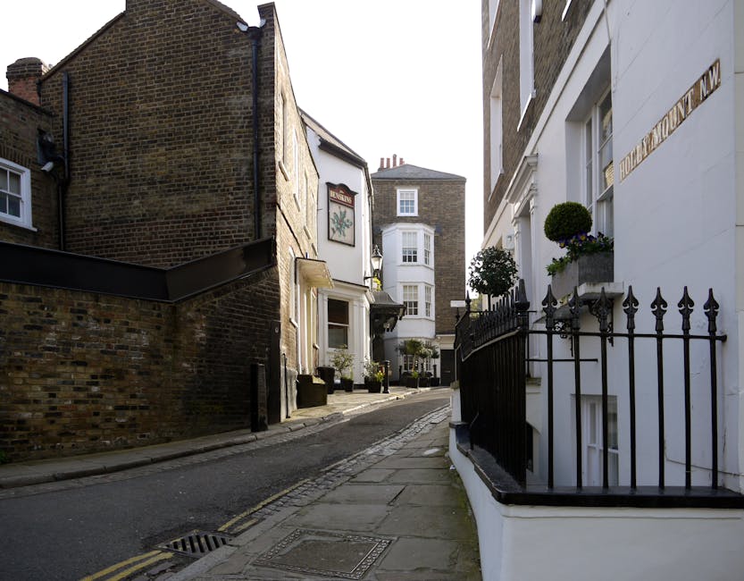 A quiet residential street in Beddington with a gently curving asphalt road, lined by a mix of terraced and semi-detached houses with white, red brick, and painted facades. Some houses display potted plants near doorways and small front gardens. The pavement on the right side has evenly spaced concrete slabs, and a black metal bollard is visible along the sidewalk. In the background, a historic church with a prominent tower and battlements stands at the end of the street under a bright blue sky with scattered clouds. The scene is well-lit and appears to be daytime. This setting reflects a typical UK suburban area suitable for home relocation and furniture transport, often serviced by companies like Man with Van Beddington during moves involving packing and loading processes.
