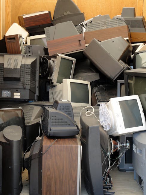 A large pile of old electronic waste and vintage computer equipment indoors, including CRT monitors, desktop computers, and old telephones, stacked on top of each other in a cluttered manner, with some items leaning against a wooden wall. The devices are mostly grey and beige with visible cables and power cords tangled among them. A few cardboard boxes with wooden-veneered exteriors are also part of the collection, indicating disorganized disposal or recycling of outdated technology. This scene illustrates the collection and removal process of bulky waste items as part of a house relocation or clearance service, such as offered by Man with Van Beddington, which includes handling and transporting electronic and household waste efficiently.