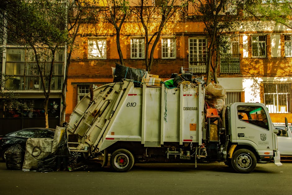 A white waste collection truck parked on a street in Beddington during daytime, loaded with various bulky waste items including large black garbage bags, cardboard boxes, and an assortment of household disposal items. The truck is positioned in front of a multi-storey residential building with orange brick walls and multiple windows, some of which have curtains, while trees with green leaves partially shade the scene. The truck belongs to a waste management service and is involved in the collection of bulky household waste, possibly as part of a home relocation or removal process managed by Man with Van Beddington. The loading area of the truck is filled with assorted waste, and a trolley and straps are visible, indicating an active loading process. The environment shows clear daylight, with shadows cast by the trees, and the scene captures essential steps in waste clearance related to house removals and moving services.