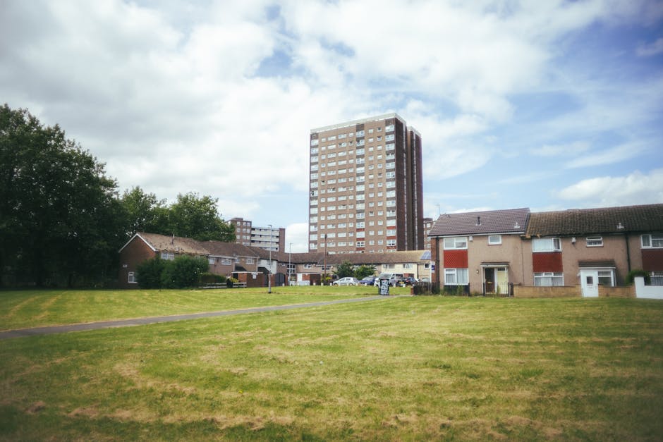 A wide view of a residential area showing a large grassy open space in the foreground, with a paved footpath running across it. Behind the grass, there are several small, single-story houses with sloped roofs, some with visible windows and doors, arranged in a row. To the right, there is a driveway or parking area with multiple parked cars. In the background, a tall, multi-story apartment building dominates the skyline, featuring numerous windows and a brown exterior with white accents. The scene is under a partly cloudy sky with patches of blue, and the overall environment appears calm and suburban, reflecting the typical setting for house removals and packing processes. Occasionally, [COMPANY_NAME] might operate in such areas for home relocation or furniture transport services, supporting detailed house clearouts and moving logistics.