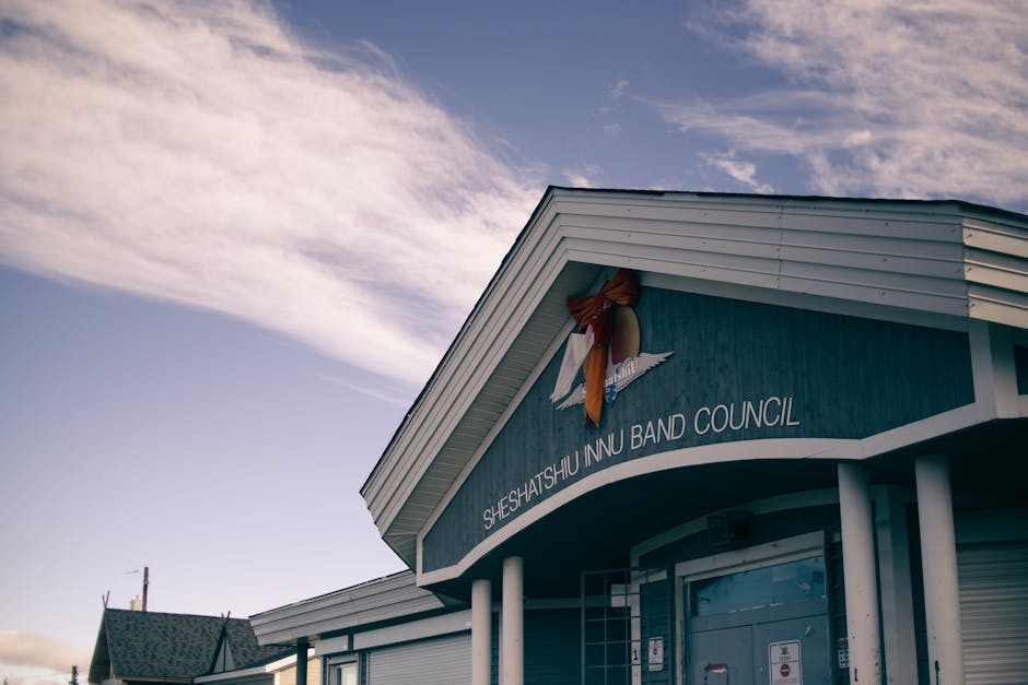 The exterior of a building identified as Sheshatshu Inn Nu Band Council, featuring a blue facade with white trim and a prominent curved entrance canopy supported by white columns. A decorative ribbon with a bow is attached near the roofline above the signage. The building is set against a partly cloudy sky with soft daylight illuminating the scene. Nearby, there are rooftops of other structures, indicating a small town or community setting. This scene relates to house removals and moving logistics, where careful packing, loading, and transportation of furniture and boxes are essential steps handled by companies like Man with Van Beddington during local relocations within Sutton or Beddington areas.