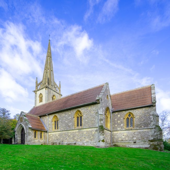 Exterior view of a historic stone church with a tall, pointed spire and Gothic-style arched windows, situated on a grassy area under a bright blue sky with scattered clouds. The church features a sloped roof covered with reddish-brown tiles and a small stone porch at the entrance. The surrounding environment includes a well-maintained lawn and some trees in the background. This image relates to house removals and moving services provided by Man with Van Beddington, illustrating an outdoor setting commonly associated with relocation logistics and the transport of household furniture and items.