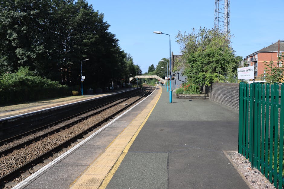 A clear view of Hackbridge station platform showing railway tracks running parallel along the left side, with a yellow safety line adjacent to the platform edge. The platform surface is paved with dark grey concrete, featuring tactile paving strips near the edge for visually impaired passengers. Several blue-painted poles with lamps extend along the platform, and a small, white signboard indicating station information is visible on the right side, next to a green metal fence that encloses a small garden area with bushes and a tree. In the background, there are residential and commercial buildings, including a brick structure and an overhead bridge crossing over the tracks, under a bright, clear sky. The station appears tidy and well-maintained, suitable for home relocation and furniture transport services by [COMPANY_NAME], with space for moving vans and loading activities happening on the platform exterior.