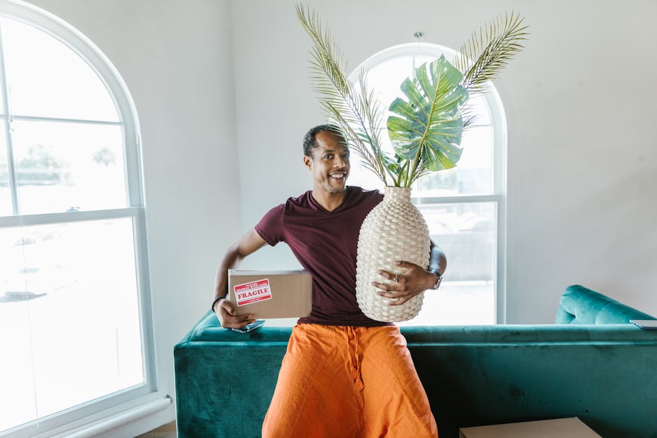 A man with dark skin and short curly hair is inside a bright room with natural light coming through large arched windows. He is smiling and holding a large white textured ceramic vase filled with green tropical leaves and palm fronds, supporting the base with one arm. In his other hand, he grasps a medium-sized cardboard box marked with a red 'FRAGILE' sticker. The man is dressed in an orange pair of trousers and a maroon t-shirt. Behind him is a teal upholstered sofa against the wall, with some additional cardboard boxes stacked on a side table. The room has a light-colored wall and the window frames are white. This scene illustrates the packing and home relocation process, with the man preparing items for the move, consistent with services offered by Man with Van Beddington during house removals and furniture transport.