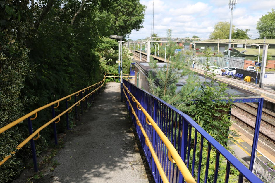 A clear view of Hackbridge station platform showing railway tracks running parallel along the left side, with a yellow safety line adjacent to the platform edge. The platform surface is paved with dark grey concrete, featuring tactile paving strips near the edge for visually impaired passengers. Several blue-painted poles with lamps extend along the platform, and a small, white signboard indicating station information is visible on the right side, next to a green metal fence that encloses a small garden area with bushes and a tree. In the background, there are residential and commercial buildings, including a brick structure and an overhead bridge crossing over the tracks, under a bright, clear sky. The station appears tidy and well-maintained, suitable for home relocation and furniture transport services by [COMPANY_NAME], with space for moving vans and loading activities happening on the platform exterior.