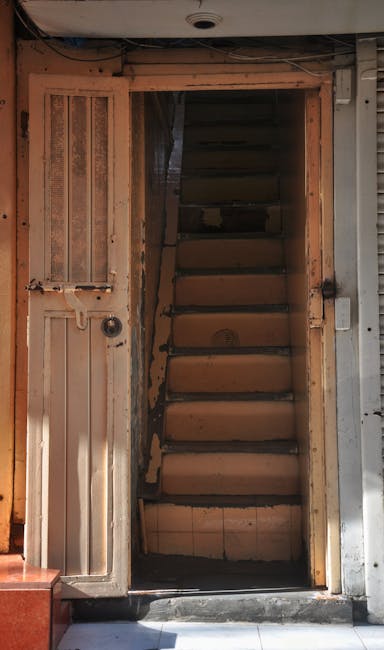 A narrow wooden staircase inside a property, with six steps visible leading upwards, bordered on the left by a wooden door with a metal handle and keyhole. The surrounding walls consist of unfinished wooden framing, showing exposed wood and some insulation material. The staircase appears aged, with darkened, slightly worn treads. The area is lit by natural light coming from the front, casting soft shadows. To the right of the doorway, part of a metallic structure, possibly part of a construction or renovation setup, is visible. The ground floor features a dark, narrow opening, consistent with a small entrance or access point, likely part of a home relocation or packing process, as handled by Man with Van Beddington, within the context of delicate furniture transport or moving logistics involving fragile items and stairwell navigation.
