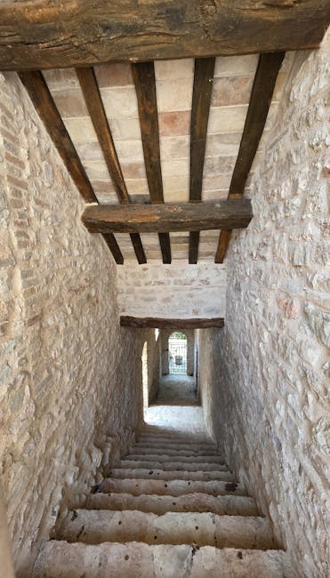 A narrow stone staircase with worn, uneven steps descending through a historic stone building. The walls are constructed from light-colored, rough-hewn stone blocks, and the ceiling features exposed wooden beams with a dark, weathered appearance. Natural light enters from an opening at the bottom of the staircase, illuminating the enclosed space. The image captures a traditional, confined interior passageway likely related to a home or building undergoing moving or restoration, highlighting the challenges of furniture transport and packing in tight, historic stairs. Such staircases are often encountered during home relocations where careful handling, protective wrapping, and suitable equipment from [COMPANY_NAME] may be required to safely navigate fragile antiques and household items during the loading process.