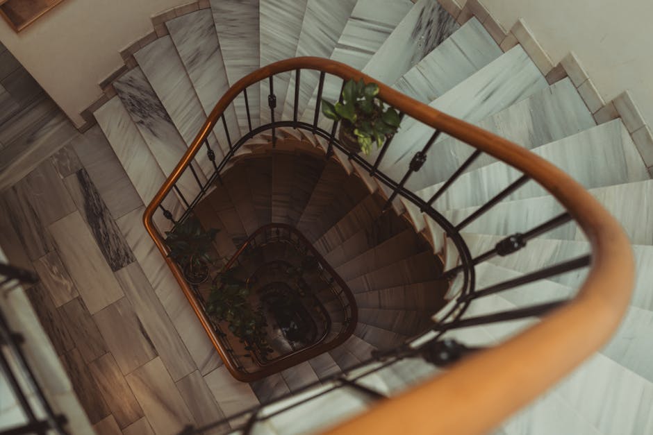 A narrow wooden staircase inside a property, with six steps visible leading upwards, bordered on the left by a wooden door with a metal handle and keyhole. The surrounding walls consist of unfinished wooden framing, showing exposed wood and some insulation material. The staircase appears aged, with darkened, slightly worn treads. The area is lit by natural light coming from the front, casting soft shadows. To the right of the doorway, part of a metallic structure, possibly part of a construction or renovation setup, is visible. The ground floor features a dark, narrow opening, consistent with a small entrance or access point, likely part of a home relocation or packing process, as handled by Man with Van Beddington, within the context of delicate furniture transport or moving logistics involving fragile items and stairwell navigation.
