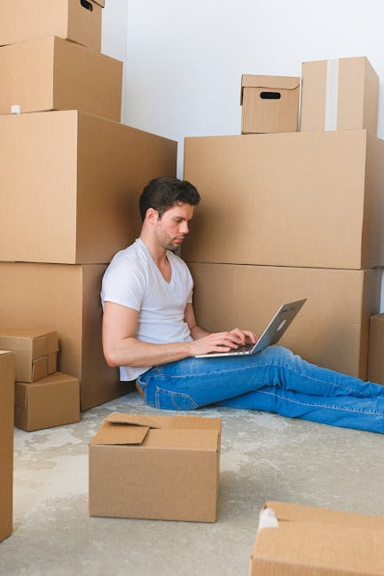 A man with short dark hair and light skin, wearing a white t-shirt and blue jeans, is sitting on the floor inside a room surrounded by numerous cardboard boxes of various sizes, some sealed with packi