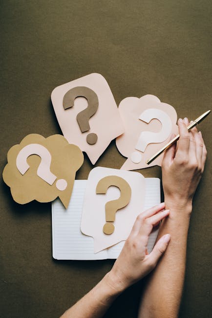 A person is seated at a table, holding a pen in their right hand and resting their left hand on a notebook. Surrounding the hands are multiple paper cutouts shaped like speech bubbles and clouds, each featuring a large question mark in the center. These objects are made from beige and white paper, with some cutouts placed on top of a lined notepad, suggesting notes or planning related to house removals. The scene is set on a brown surface, possibly a table, with natural lighting highlighting the materials used for packing and potential moving considerations. The image visually emphasizes questions and planning involved in home relocation or furniture transport, aligning with the theme of understanding removal estimates, as presented by Man with Van Beddington.